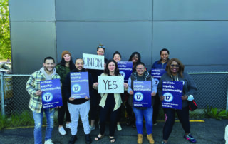 Image of new PROTEC17 members, the team at King County Regional Homelessness Authority, standing in front of a gray wall and holding celebratory union signs.