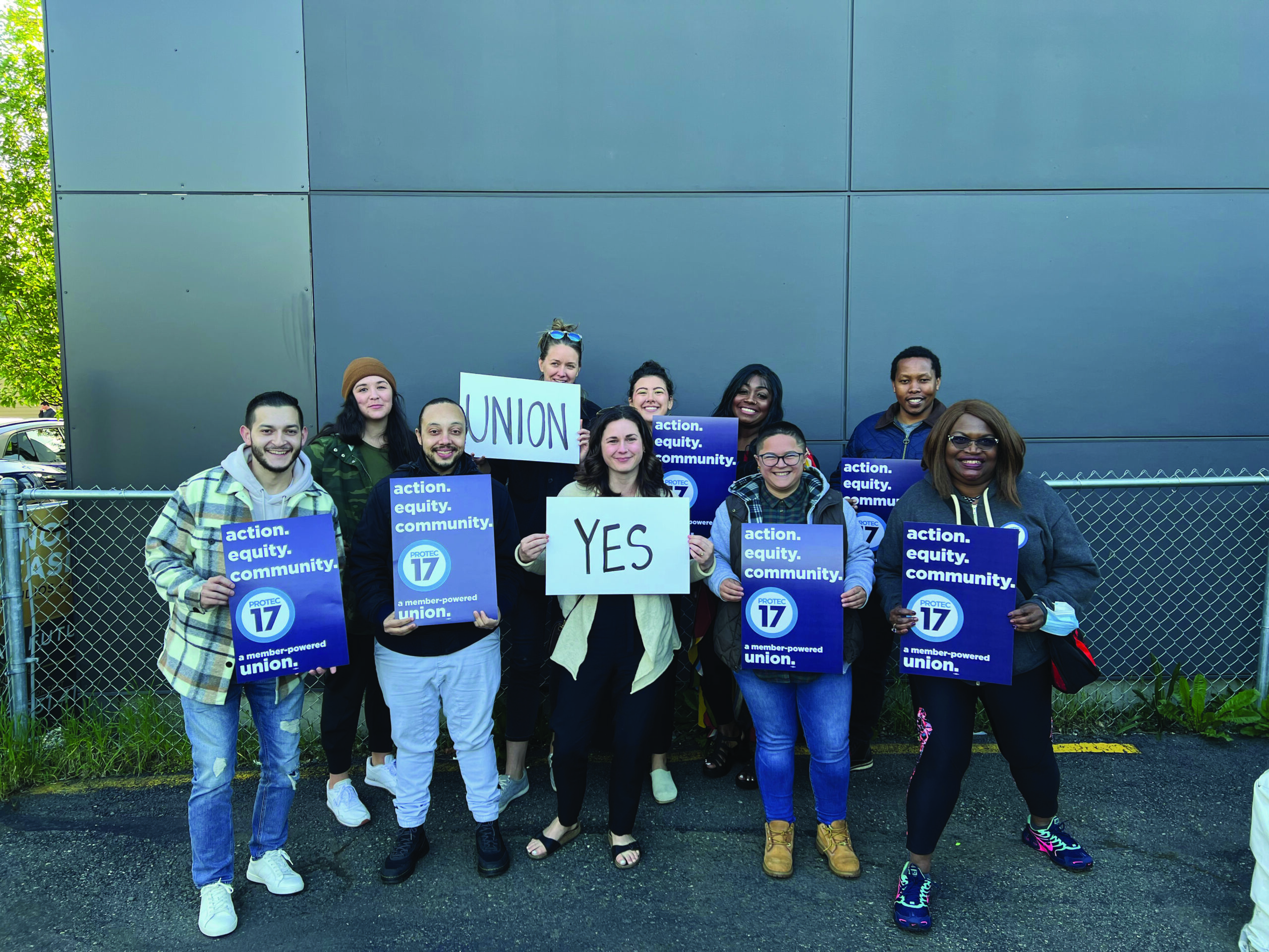 Image of new PROTEC17 members, the team at King County Regional Homelessness Authority, standing in front of a gray wall and holding celebratory union signs.