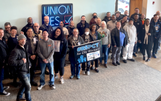 Picture of Seattle Coalition of City Unions (CCU) members and staff at the bargaining session on March 15. They are standing in front of a "Union Yes" sign and holding the #RSPCT campaign banner.