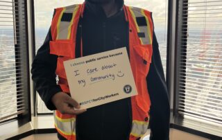 Picture of a City of Seattle worker in a bright construction vest, holding a sign that reads: "I choose public service because I care about my community :) #RSPCTForCityWorkers"