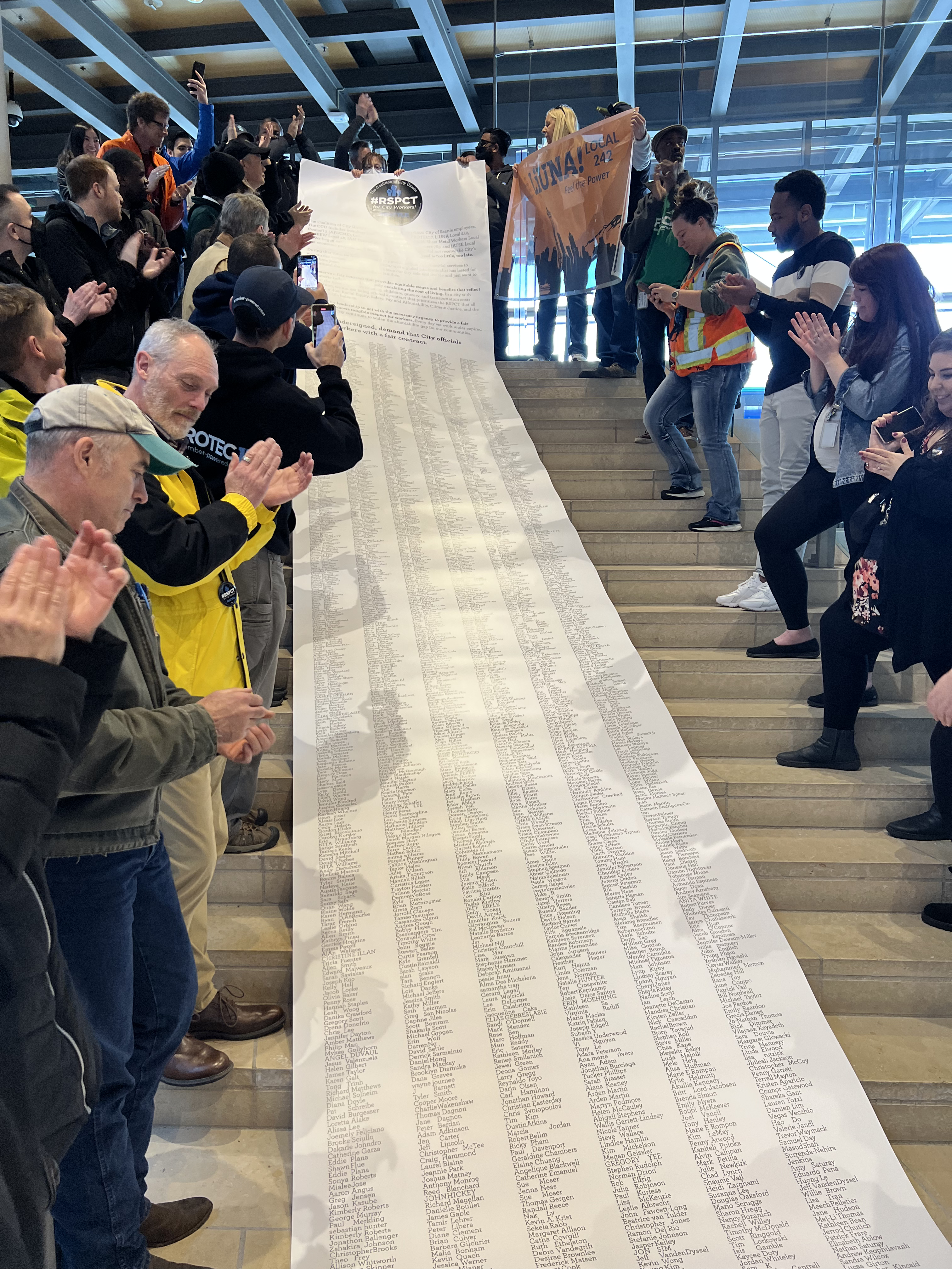 Picture of Seattle Coalition of CCU members clapping at the sight of the large scroll with petition signatures, unfurling down the steps of City Hall.