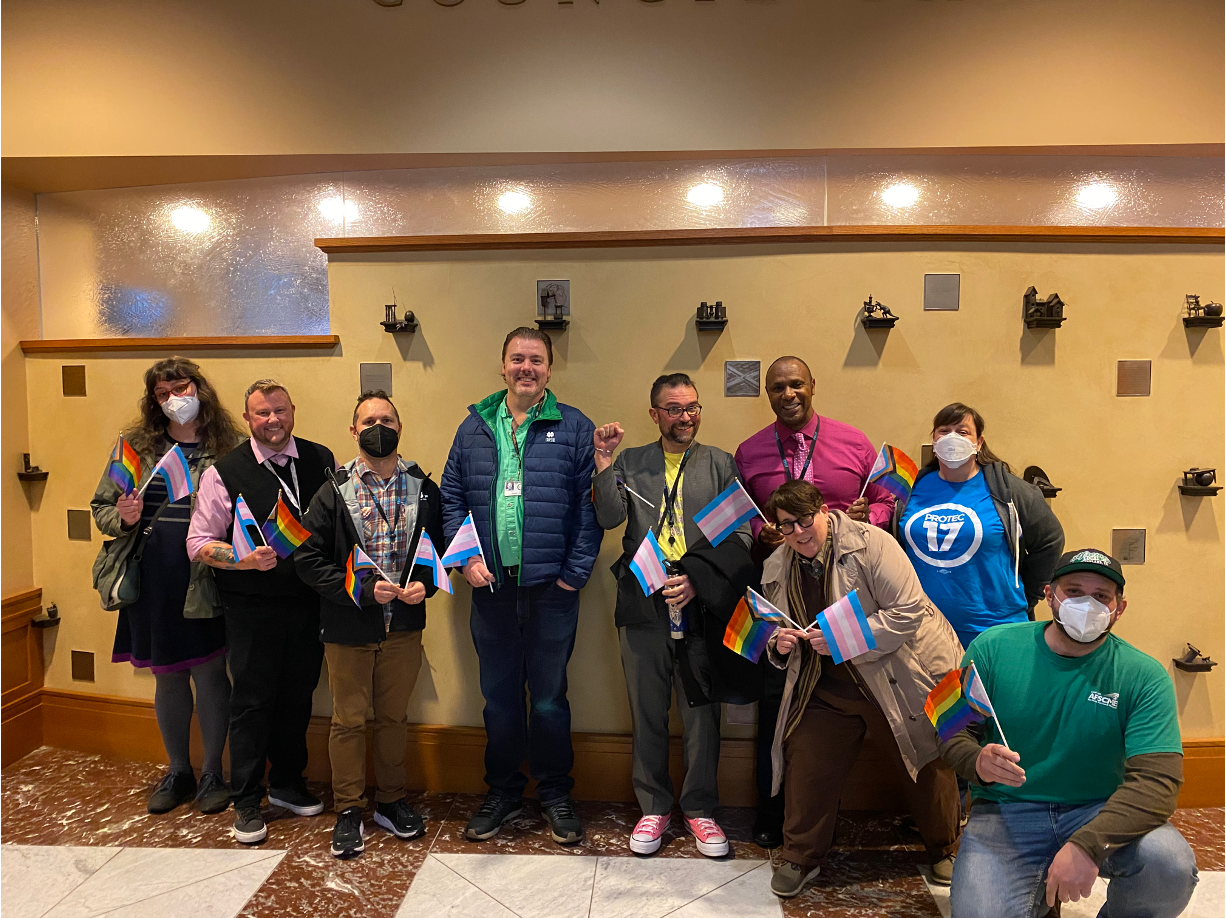 Picture of PROTEC17 Portland members holding mini trans pride and LGBTQIA+ pride flags and smiling.