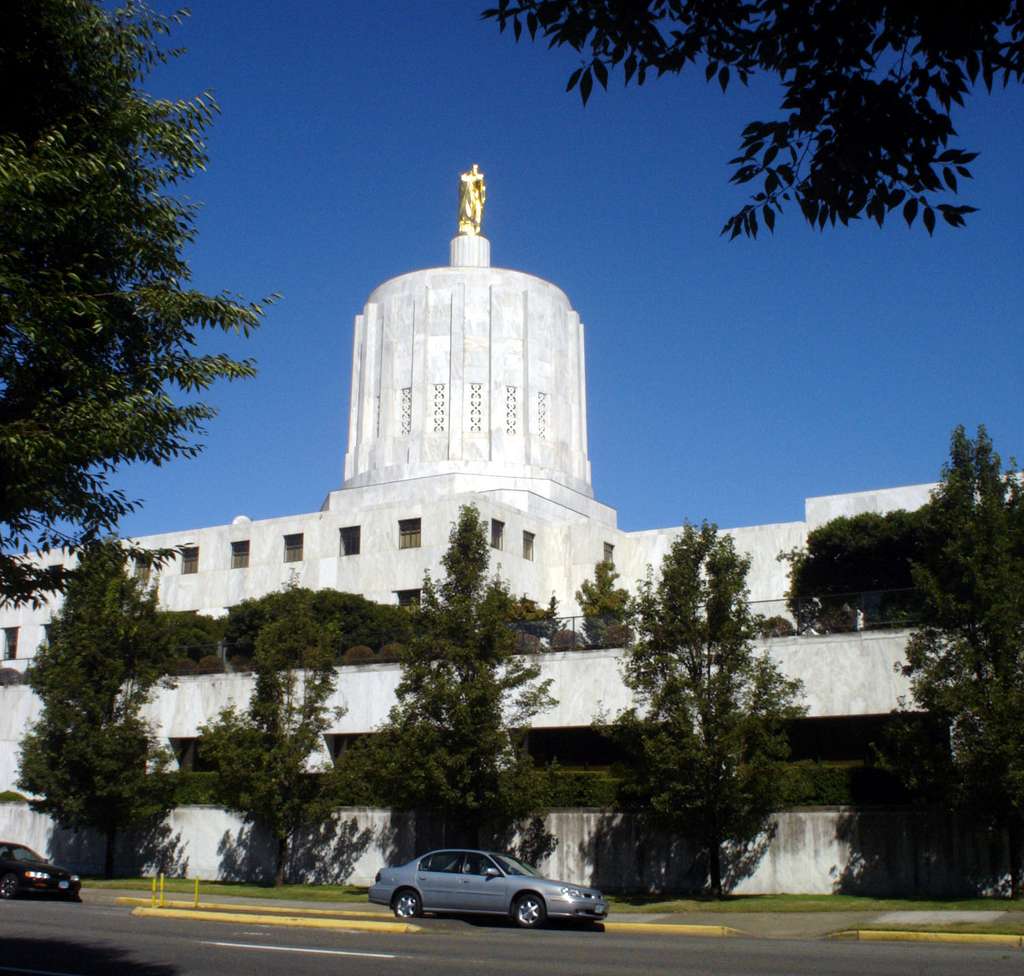 Picture of the Oregon state capitol building. The sky is blue and there are trees pictured in front of the building.