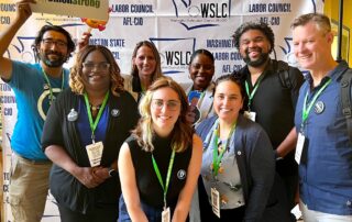 Picture of PROTEC17 members, e-board members, and staff smiling and posing in front of the WSLC backdrop. From left to right: Dan Santon, Jennell Hicks, Karen Estevenin, Brenna Stroup, Alisha Gregory-Davis, Jessica Olives, Brandon Hersey, and Mark Watson. Dan is holding a sign that reads, "#UnionStrong."