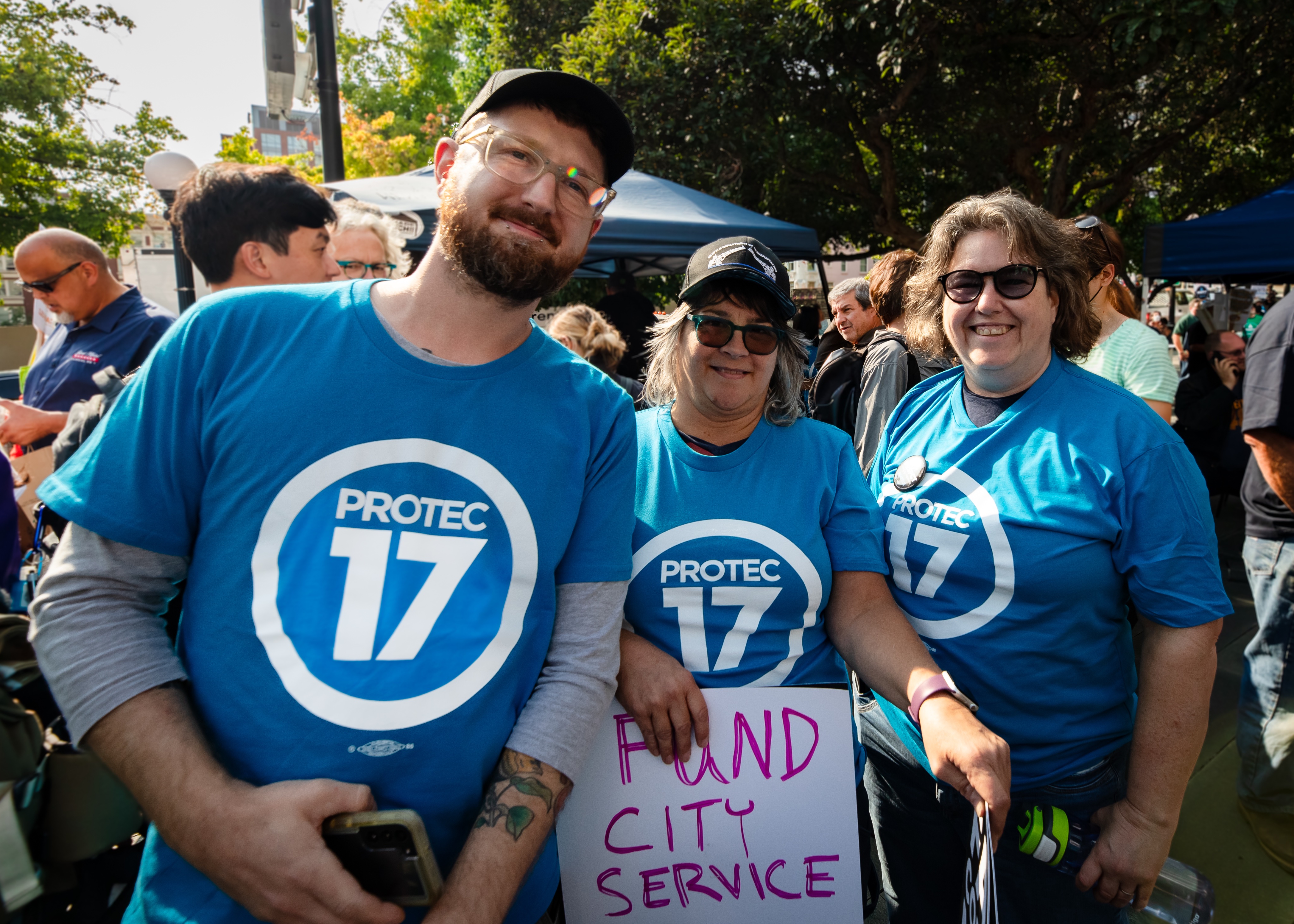 Picture of coalition members wearing PROTEC17 shirts, smiling, and holding a sign that reads, "FUND CITY SERVICE"