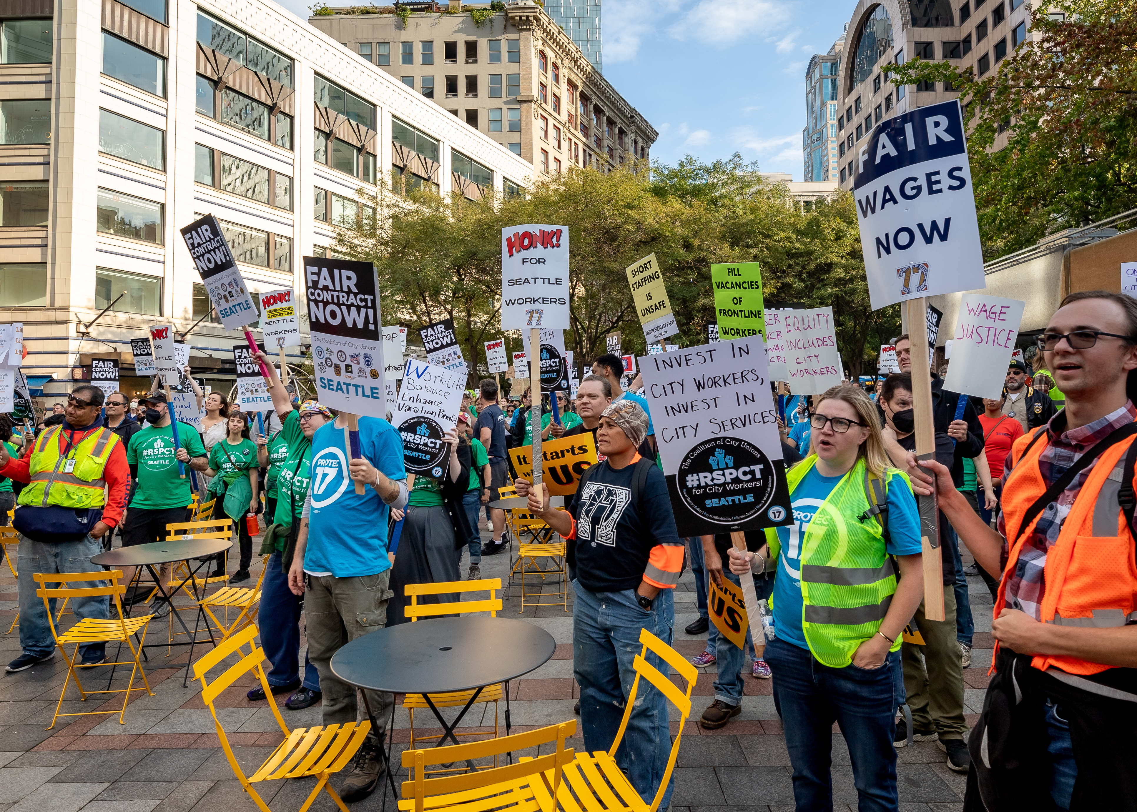 A crowd of coalition union members gather at the end of their march at Westlake Center in downtown Seattle. Folks are pictured wearing their union gear, safety vests, and holding up their signs to demand a fair contract.
