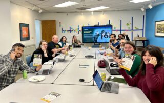 Picture of PROTEC17 members and staff at the phone banking event for Seattle City Council Candidate (District 3), Alex Hudson. Everyone is sitting around the large conference table in front of a screen with Alex's campaign imagery, smiling.