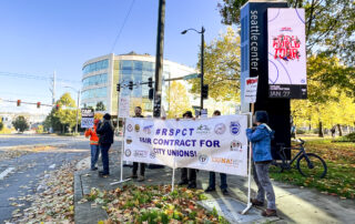 Coalition members stand together on the corner of Seattle Center holding a RSPCT campaign banner. They are standing in front of the Seattle Center sign with digital media on it.