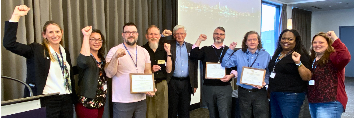 Picture of PROTEC17 Executive Director and Executive Board members with REC award winners at the Oct. 2023 REC meeting. Everyone is smiling and standing to the side of a projector screen in the background. Everyone is smiling and holding their fists up in solidarity.