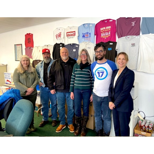 Spokane Regional Health District member Nick stands with other labor leaders as a Labor Council Delegate. He is wearing a long sleeve PROTEC17 shirt and is smiling. Everyone is standing in front of union t-shirts hung on the wall.