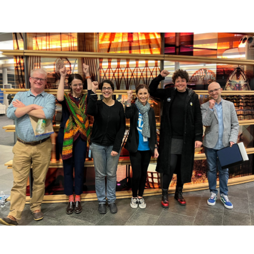 PROTEC17 Seattle Stewards stand with. Union Representative Kaite at Seattle City Hall. They are all standing in from of amber-colored glass with pictures of famous areas around Seattle. Everyone is smiling and some folks are holding up fists raised in solidarity.