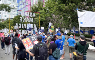 PROTEC17 members and staff march on the street alongside union siblings and community members. Everyone is wearing their union gear and/or protest wear and is walking towards the Palantir building.