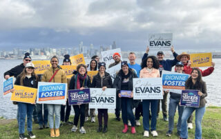 PROTEC17 members and staff stand with local, Seattle candidates running for Seattle City Council. They are standing on grass--water and the Seattle skyline are both visible in the background. Folks are holding campaign signs and wearing their PROTEC17 union gear.