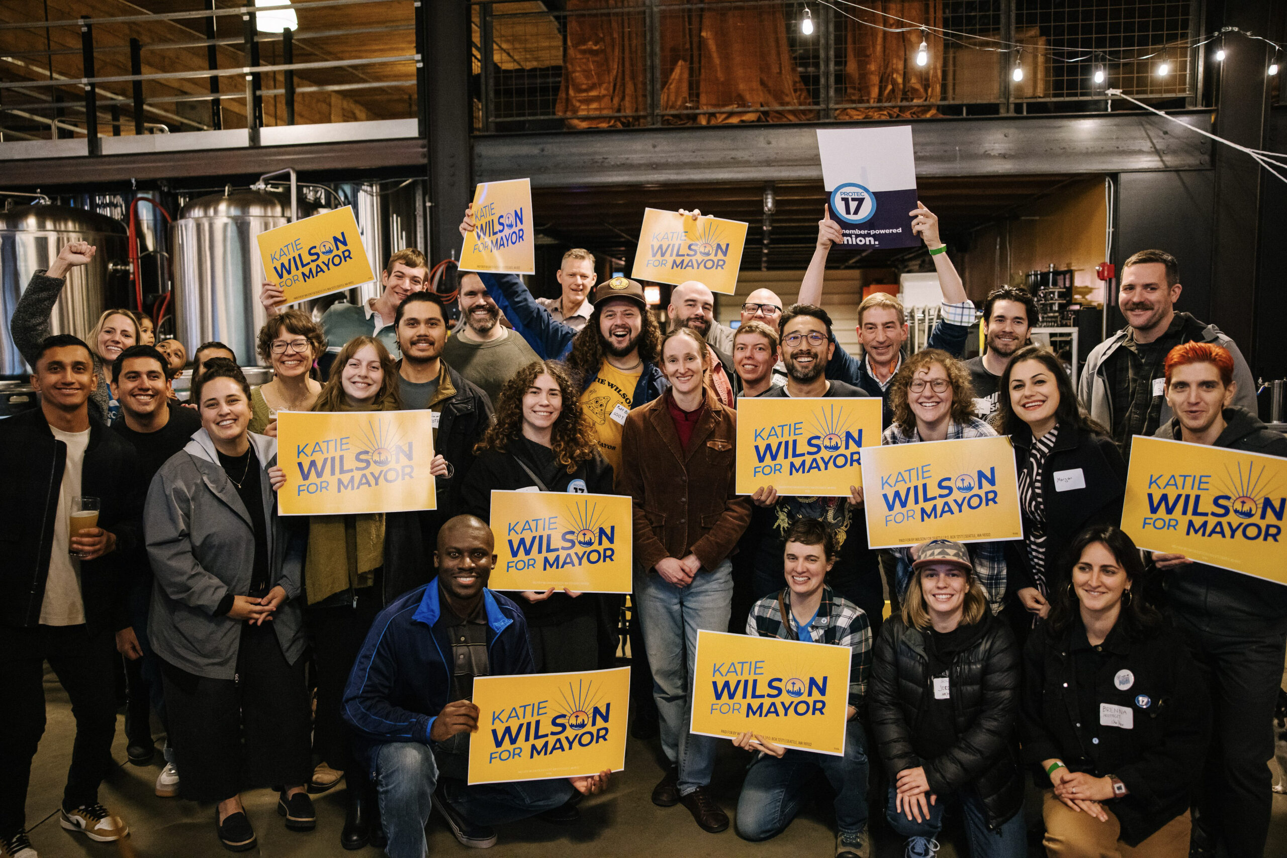 PWR! Mixer – Oct. 2025 PROTEC17 members, staff, and friends stand together for a photo. Everyone is smiling enthusiastically and holding up Kaite Wilson (mayoral candidate) campaign signs.