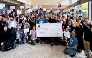 PROTEC17 members and staff stand in the lobby at the King County Chinook building. Folks are wearing their PROTEC17 union gear and raising union signs. Everyone is smiling and raising their fists in solidarity.