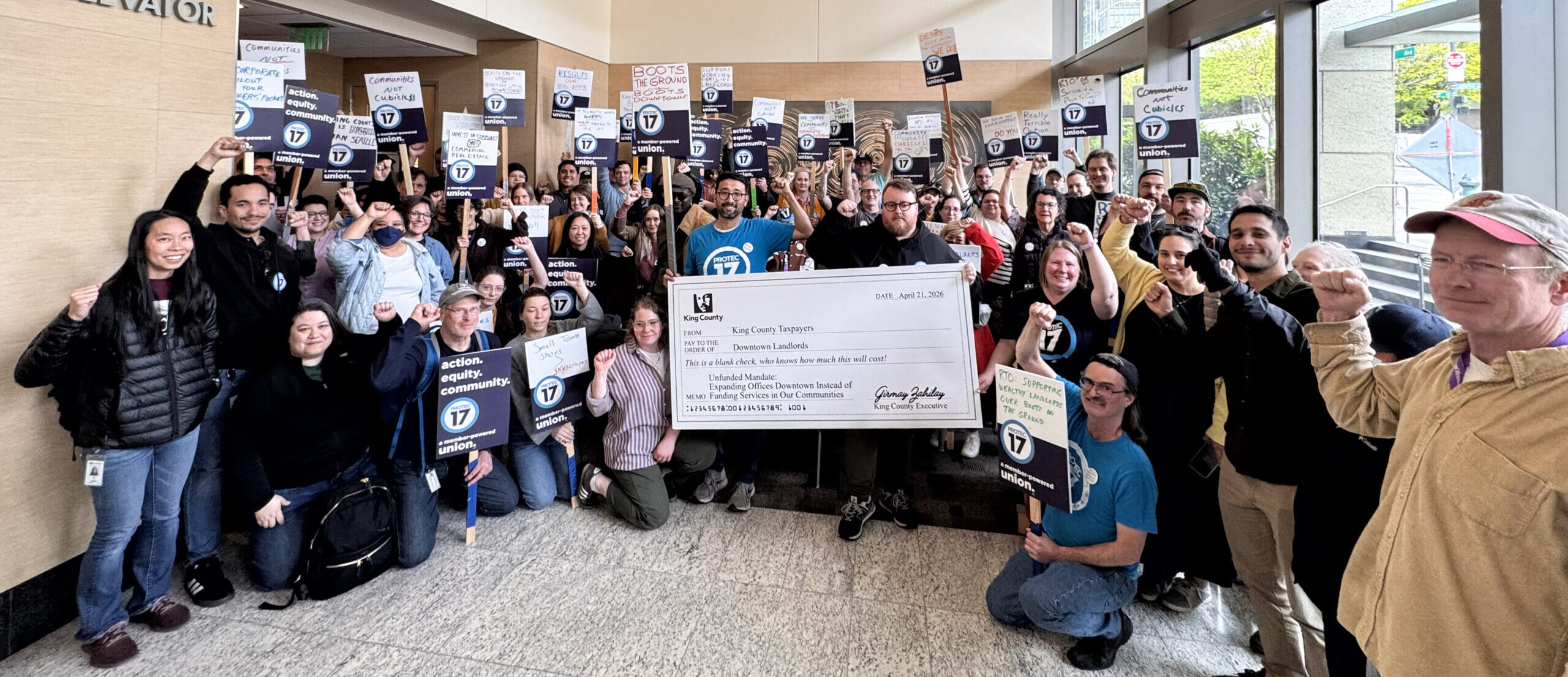 PROTEC17 members and staff stand in the lobby at the King County Chinook building. Folks are wearing their PROTEC17 union gear and raising union signs. Everyone is smiling and raising their fists in solidarity.