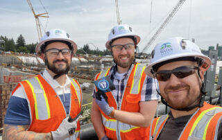 Portland Chapter members who work in the Filtration Team for the City of Portland pose together at a construction site at a future water treatment facility. They are smiling and one member is holding up the PROTEC17 logo. They are wearing hard hats and safety vests.
